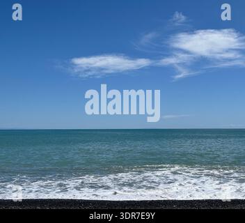 Blick auf den Horizont über ein blaues Meer vor einem hellblauen Himmel mit Whiskywolken von einem Kiesstrand Stockfoto