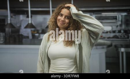Frau, die Haare mit der Hand berührt, leichtes Lächeln und geschlossene Augen in einer Restaurantküche; ruhige Besinnung. Stockfoto