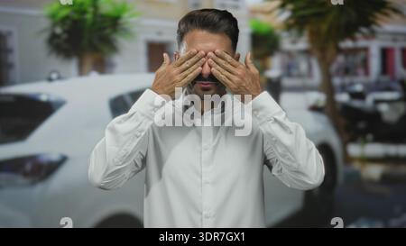 Junger hispanischer Mann mit weißem Hemd bedeckt den Mund mit beiden Händen auf der belebten Straße und sieht schockiert aus; Überraschung. Stockfoto