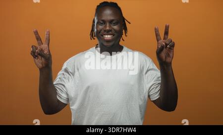 Junger afroamerikanischer Mann in weißem T-Shirt mit doppelten Friedenszeichen mit beiden Händen im Studio mit orangefarbenem Hintergrund; Glück Frieden Positivität. Stockfoto