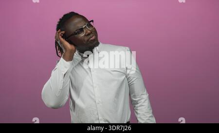 Mann, der mit der Hand das Ohr berührt und im Studio eine Brille vor rosa Hintergrund trägt; nachdenkliche Konzentration. Stockfoto