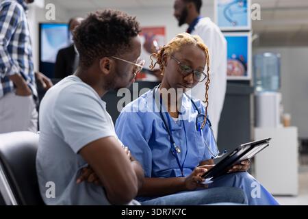Aufmerksame Krankenschwester in blauen Peelings führt männliche Individuen durch die medizinische Untersuchung und überprüft digitale Aufzeichnungen im Wartezimmer. Professionelle Ärztin bespricht Krankenversicherungsinformationen mit Patienten. Stockfoto