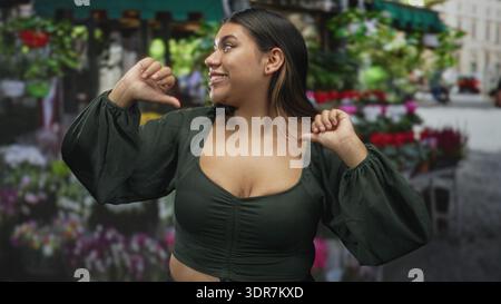 Frau zuckt mit den Händen, entblößte Brust auf einem geschäftigen Straßenblumenmarkt, während sie lächelt und sich zwischen Blumensträußen posiert; spielerische Leichtigkeit. Stockfoto