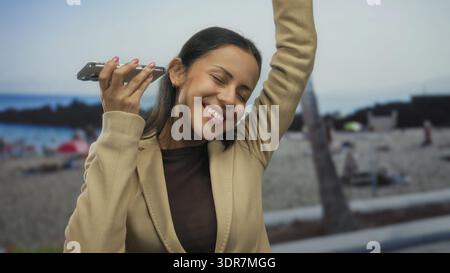 Die junge Frau tanzt fröhlich an einem sonnigen Strand, hört Musik von ihrem Handy, mit dem Meer im Hintergrund und trägt eine braune Jacke, Embodyin Stockfoto