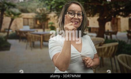 Hispanische Seniorin mit Brille zeigt mit dem Zeigefinger auf Terrassentische in einem Innenhof eines Cafés an einem Steingebäude; Vertrauensberatung. Stockfoto