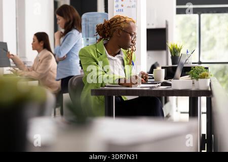 Der afroamerikanische Manager überprüft das Projekt auf dem Laptop und macht sich Notizen, um die Vorbereitung auf das bevorstehende Meeting zu gewährleisten. Engagierter Teamleiter, der die betriebliche Planung der Abteilung und die Geschäftsstrategien verwaltet. Stockfoto
