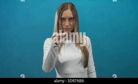 Junge hispanische Frau, die Hand zeigt, die von der Hüfte nach oben in einem Studio mit blaublauem Hintergrund und neutralem Ausdruck auf die Kamera zeigt; Entschlossenheit. Stockfoto