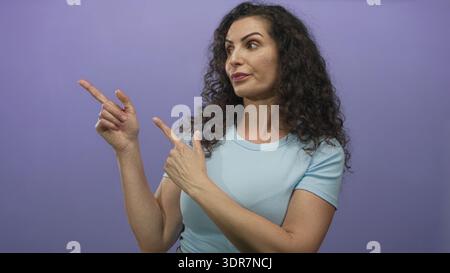 Hispanische Frau mit hellblauem Hemd zeigt beide Zeigefinger in lila Atelier nach links; Richtung. Stockfoto