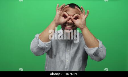 Junger hispanischer Mann in gestreiftem Hemd, der Handferngläser über lächelndem Gesicht im grünen Studio macht; verspielte Neugier. Stockfoto