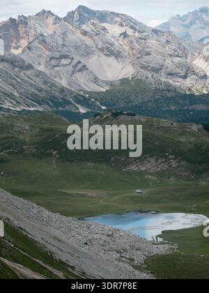 Dolomiten, Italien - 10. Juli 2025: Majestätische Bergkette mit einem ruhigen Bergsee, abgelegenen Hütten und grünen Hängen unter bewölktem Himmel. Stockfoto