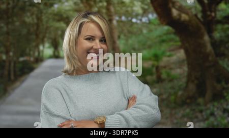Junge blonde Frau in blauem Pullover lächelt mit überkreuzten Armen auf einem gepflasterten Weg im grünen Wald; Leugnung. Stockfoto