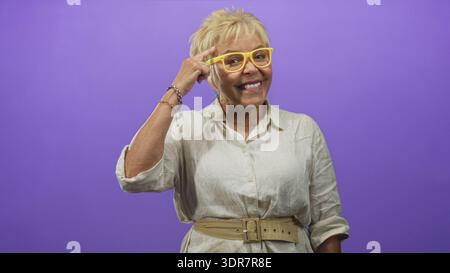 Eine ältere Frau zeigt mit dem Finger auf den Tempel, trägt gelbe Brille und beigefarbenes Leinenhemd mit Armbändern und Gürtel im Studio; Vertrauensweisheit. Stockfoto