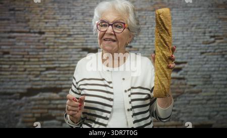 Ältere Frau, die ein langes Baguette in der rechten Hand hält, während sie mit der linken Hand in einem Studio eine Geste zur Kamera drückt; Nostalgie. Stockfoto
