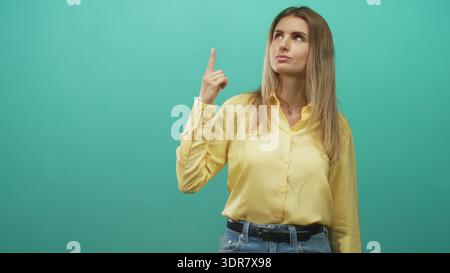 Die Frau zeigt den Zeigefinger im Studio nach oben, während sie nach oben blickt. Sie trägt gelbe Bluse und Jeans, blondes Haar sichtbar; Neugier auf Ideen. Stockfoto