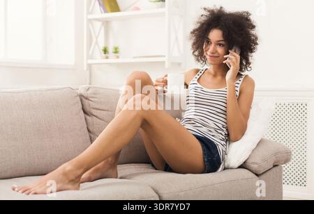 Frau, die auf einer Couch sitzt und am Telefon spricht und in ihrem Wohnzimmer einen Becher hält Stockfoto