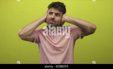 Der junge hispanische Mann bedeckt die Ohren mit beiden Händen in einem leuchtend grünen Studio-Set, während er unangenehm grimmt; Ärger. Stockfoto