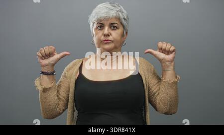 Frau zeigt in einem grauen Studio die Daumen auf die Brust, trägt Tanktop und Strickjacke mit Basketball und Armbändern; Selbstvertrauen. Stockfoto