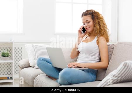 Frau, die mit Laptop auf der Couch sitzt und in einem hellen Wohnzimmer telefoniert Stockfoto