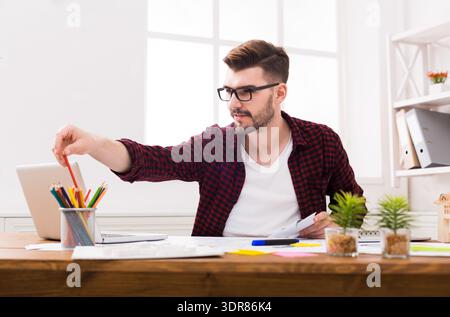 Mann, der am Schreibtisch in einem hellen Raum mit Laptop und farbenfroher Schreibwaren arbeitet Stockfoto