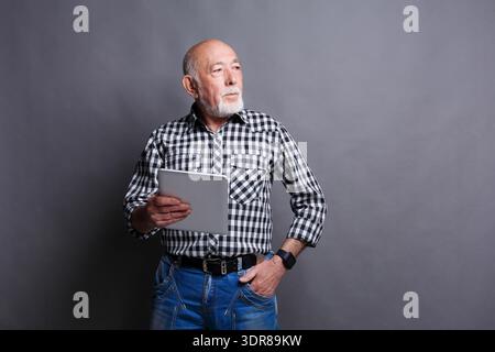 Der ältere Mann hält das Tablet und steht im Innenbereich vor grauem Hintergrund Stockfoto