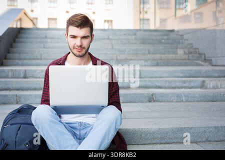 Junger Mann arbeitet am Laptop, während er auf Stufen in einem Außenbereich sitzt Stockfoto