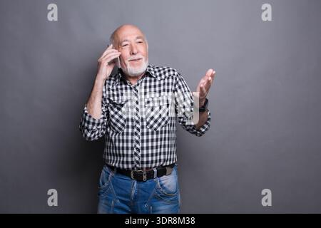 Ein älterer Mann mit Bart hält ein Telefon am Ohr, während er ein kariertes Hemd und Jeans trägt Stockfoto