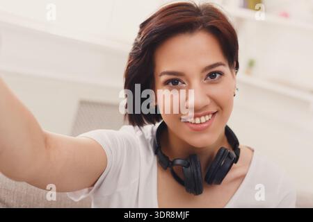 Junge Frau lächelt, während sie ein Selfie mit Kopfhörern in einem gemütlichen Innenraum macht Stockfoto