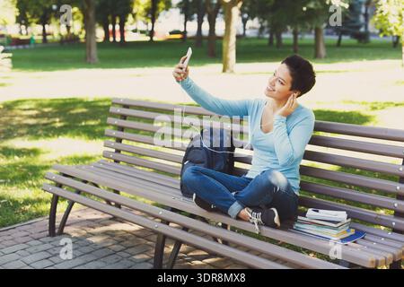 Eine junge Frau nimmt Selfie, während sie während des Sonnentages auf der Bank sitzt und Bücher im Park hält Stockfoto