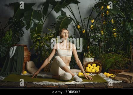 Eine Frau sitzt auf einer Yogamatte und übt eine Pose in einem Garten voller Pflanzen und Körbe mit Zitronen Stockfoto