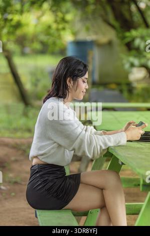Junge Frau, die ihr Smartphone benutzt, während sie an einem grünen Picknicktisch im Freien in einer natürlichen Umgebung sitzt Stockfoto