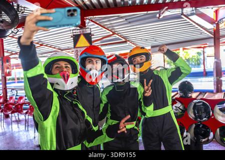 Eine Gruppe von vier verschiedenen Freunden, die bunte Helme und Rennanzüge tragen, lächeln und für ein fröhliches Selfie auf einer Gokart-Bahn posieren Stockfoto