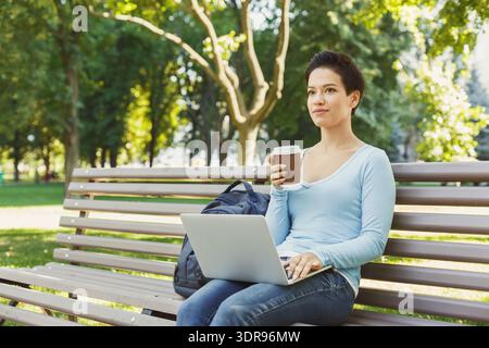 Frau mit Laptop und Kaffee sitzt am sonnigen Tag auf der Bank im Stadtpark Stockfoto