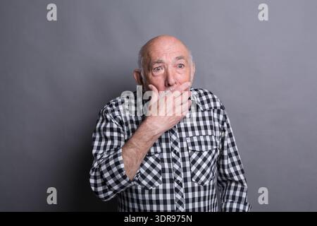 Senior man zeigt Überraschung, während er sich vor grauem Hintergrund in Studio-Setting posiert Stockfoto