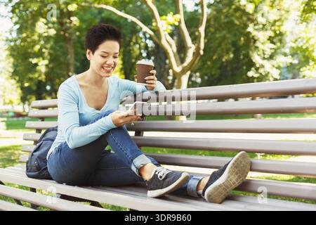 Frau genießt Kaffee und ruft das Telefon, während sie sich während des Sonnentages auf der Parkbank entspannt Stockfoto
