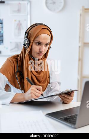 Junge muslimische Ärztin, die mit dem Patienten per Videoanruf, mit Headset und Laptop in der Klinik plaudert. Therapeut, der Empfehlungen gibt Stockfoto