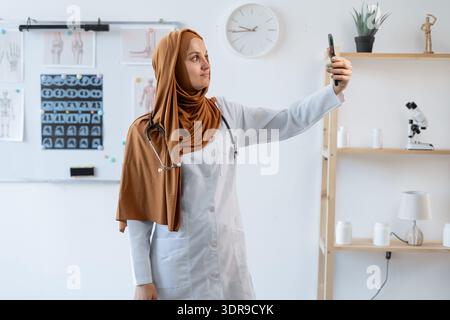 Lächelnde muslimische Ärztin im Hijab beim Selfie auf Smartphone in der Arztpraxis. Stockfoto