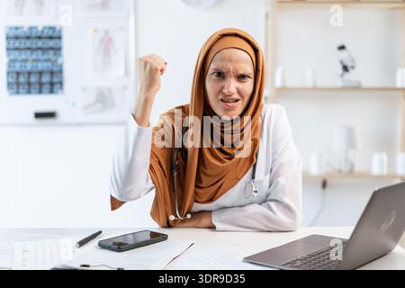 Beruflicher arabischer Allgemeinarzt. Wütende muslimische Ärztin im Hijab mit Faust-up-Geste mit aggressivem Gesicht in der Klinik Stockfoto