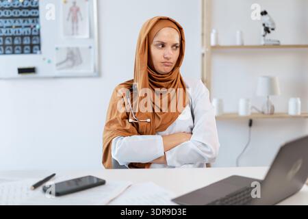 Beruflicher arabischer Allgemeinarzt. Ressentiment muslimische Ärztin im Hijab überkreuzte die Arme, während sie in der Klinik auf dem Tisch saß. Medizin Stockfoto