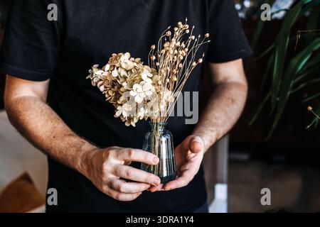 Die Person hält eine kleine Glasvase mit getrockneten Hortensien und hält sie gegen ein schwarzes Hemd. Verfaulende florale Schönheit, getrocknete Blumenküche, Schönheit in Stockfoto