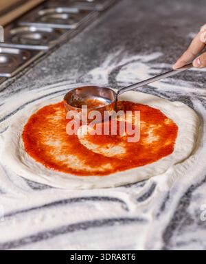 Chef Hand verteilt pasteurisierte Tomatenpaste auf eine Pizzabasis. Der Prozess der Zubereitung von Pizza. Der Koch gießt Tomatensauce über den gerollten Teig Stockfoto
