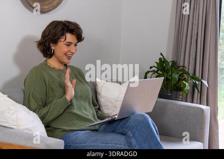 Frau, die auf einem hellgrauen Sofa im Wohnzimmer sitzt, einen grünen Pullover trägt und mit silbernem Laptop winkt Stockfoto