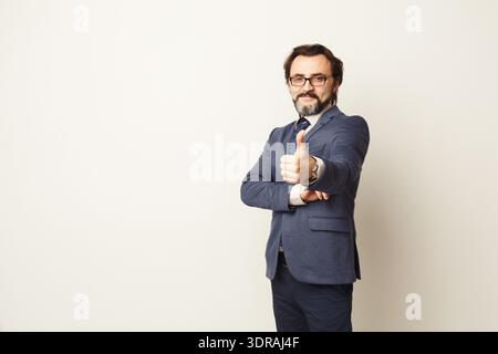 Mann im formellen Anzug, der eine Daumen-hoch-Geste mit einem selbstbewussten Ausdruck in einer einfachen Umgebung macht Stockfoto