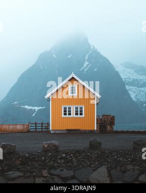 Der Blick auf ein markantes gelbes Haus erhebt sich vor dem Hintergrund einer gewaltigen, nebelummantelten Bergkette und schafft einen fesselnden Kontrast: Reine Stockfoto