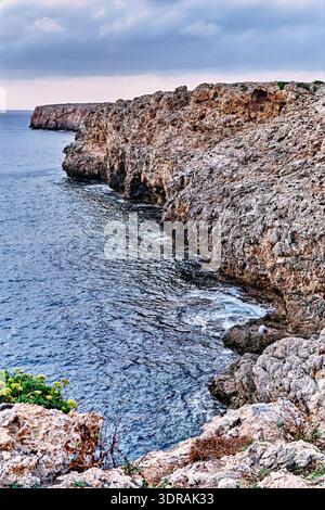 Weitläufiger Blick auf die Küste von Menorca mit zerklüfteten Felsklippen, die in das tiefblaue Mittelmeer stürzen. Wellen schlagen sanft gegen das Ufer Stockfoto