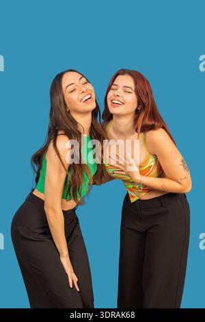 Zwei junge Frauen lachen mit geschlossenen Augen, zeigen Glück und emotionale Verbindung auf blauem Hintergrund Stockfoto