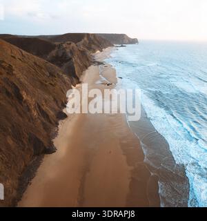 Aus der Vogelperspektive auf die zerklüftete Küste, wo die Klippen auf die Sandküste treffen. Der Kontrast aus warmen Erdtönen und kühlem Blau sorgt für eine fesselnde Szene Stockfoto