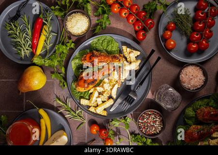 Dinner Flat Lay mit gebratenen Hähnchenkeulen mit Pommes frites auf frischem Salat, umgeben von Kirschtomaten, Rosmarin, Petersilie und verschiedenen spi Stockfoto