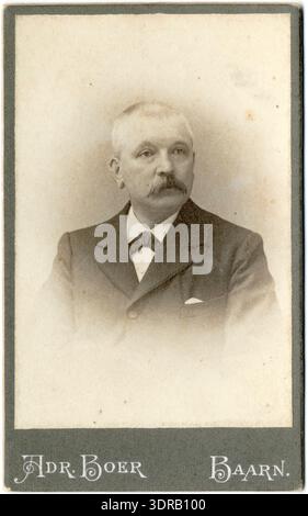 Dieses Close-up-Studio-Porträt von Adriaan Boer aus dem Jahr 1897-1912 zeigt einen älteren Holländer mit Schnurrbart, der einen Choker und eine Jacke mit einem Taschentuch trägt. Die Carte de Visite betont Gesichtszüge, Körperhaltung, Kleidung und die formale Studiokomposition, die für Baarn im späten 19. Und frühen 20. Jahrhundert typisch ist. Stockfoto