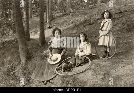 Dieses Foto von Adriaan Boer, aufgenommen zwischen 1897 und 1912 in der Nähe von Baarn, Niederlande, zeigt eine Frau mit zwei Mädchen auf unebenem Waldgelände. Die Frau trägt einen langen glänzenden Rock, eine karierte Bluse mit hohem Kragen und hält einen runden Korbmütze auf ihrem Schoß. Die beiden Mädchen tragen Röcke und Blusen mit Seemannskragen und halten Basketball in der Hand. Das Bild zeigt kindgerechte Nachbildungen und typische niederländische Kleidung aus dem späten 19. Bis frühen 20. Jahrhundert in einer Waldlandschaft. Stockfoto