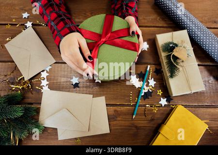 Nahaufnahme junger dunkelhäutiger weiblicher Hände mit ordentlicher Maniküre, die Neujahrsgeschenk auf Holztisch halten. Draufsicht. Frau bindet rot b Stockfoto